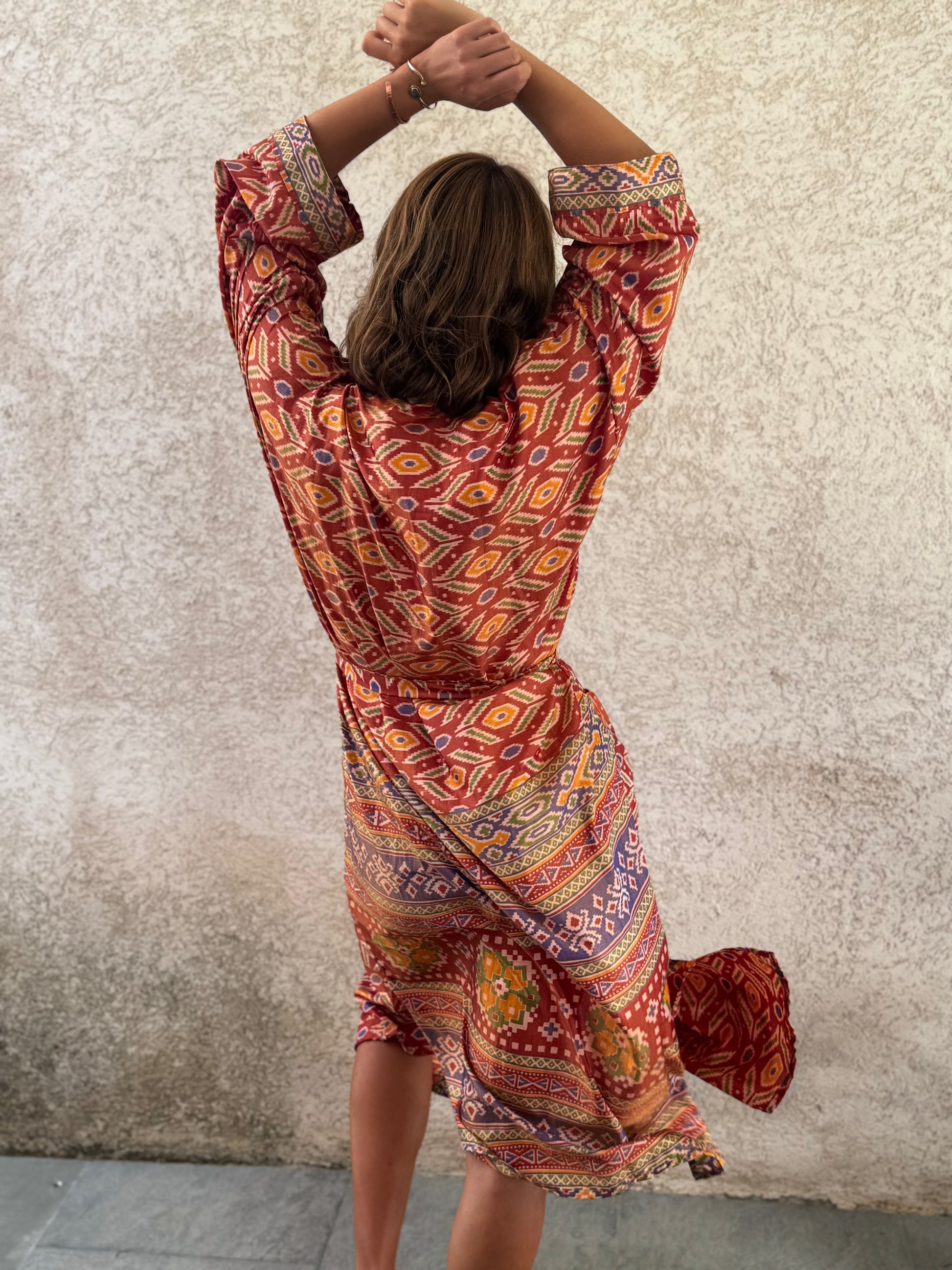 A woman viewed from behind wearing a loose, long-sleeve wrap-style dress with a colorful geometric and tribal-inspired print in orange, red, yellow, and blue, standing against a textured beige wall.