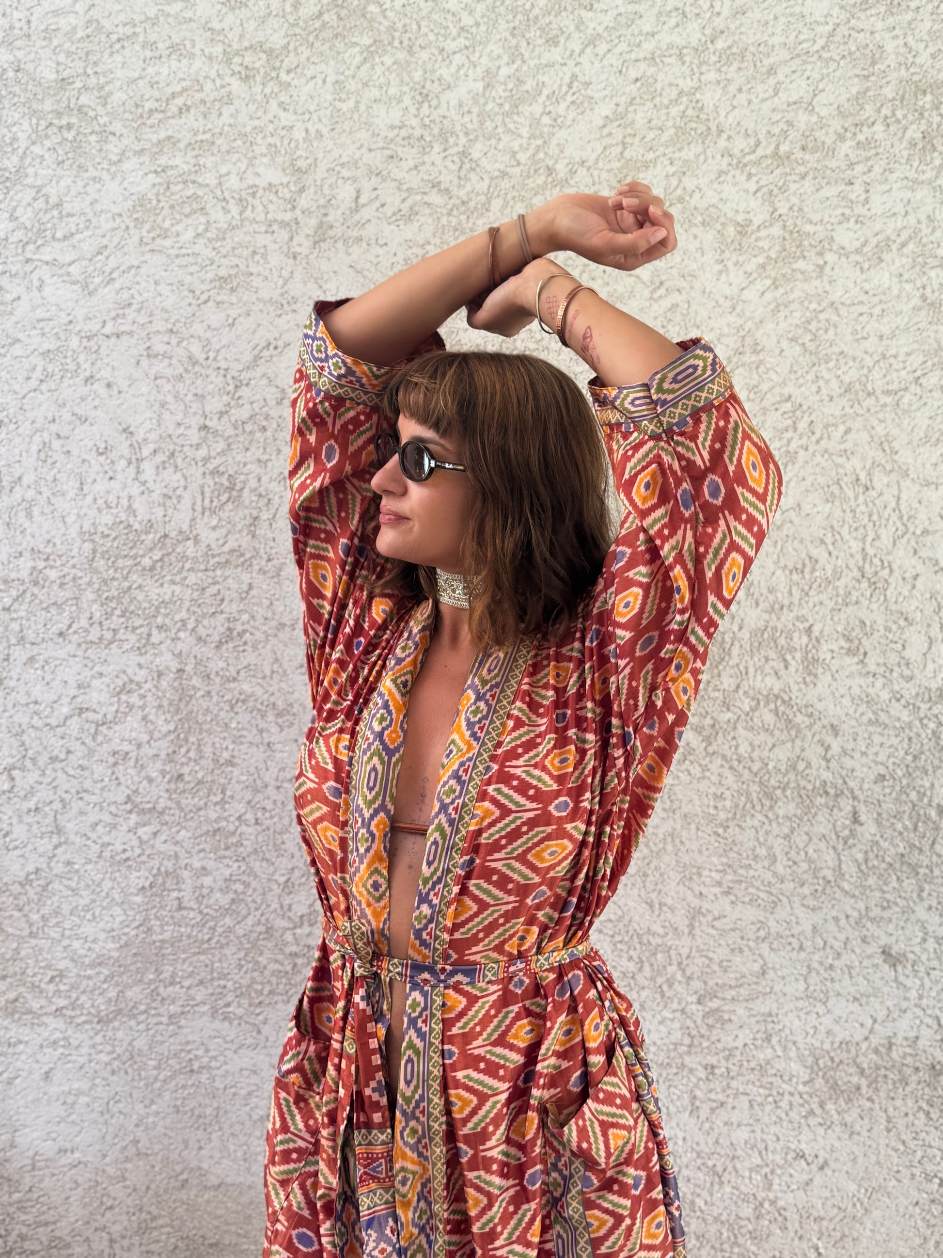A woman wearing a long-sleeve bohemian-style maxi dress with a red, orange, and yellow geometric pattern, posing with her arms raised against a textured beige wall.