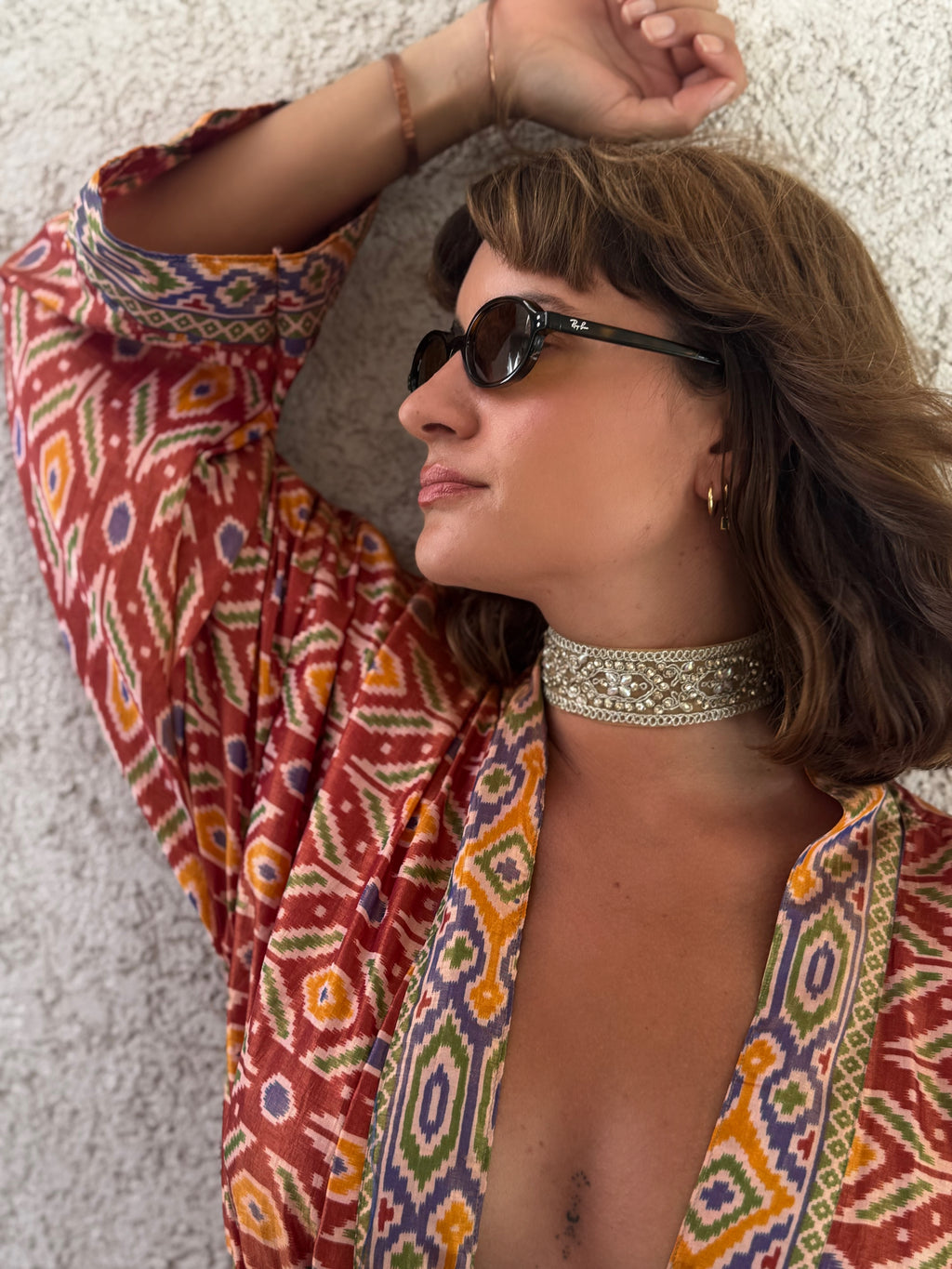 A woman wearing a red bohemian patterned kaftan top, a silver beaded choker, and black sunglasses, posing against a textured white wall.