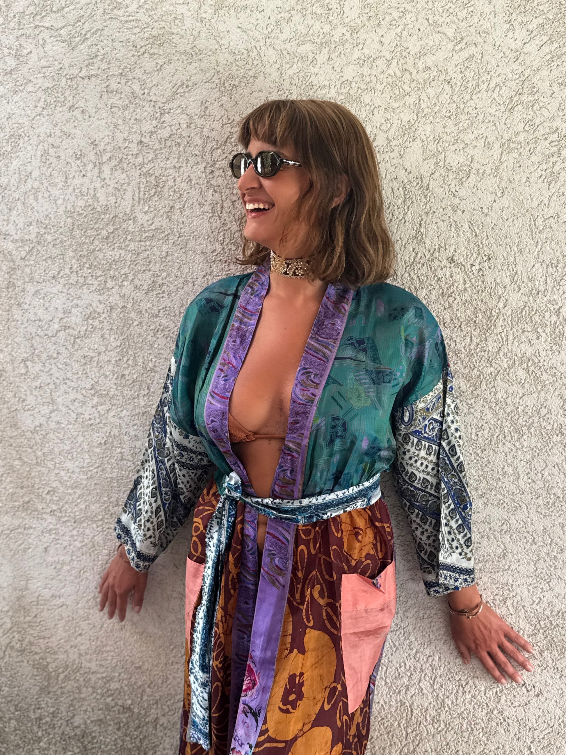 A woman wearing a colorful, patterned bohemian-style robe with purple, teal, and brown tones, standing against a textured beige stucco wall.