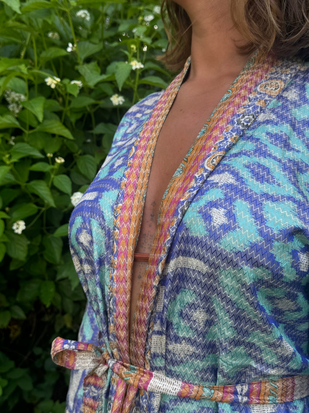 A close-up of a woman wearing a colorful patterned wrap robe with a tie waist, set against a background of green foliage and small white flowers.