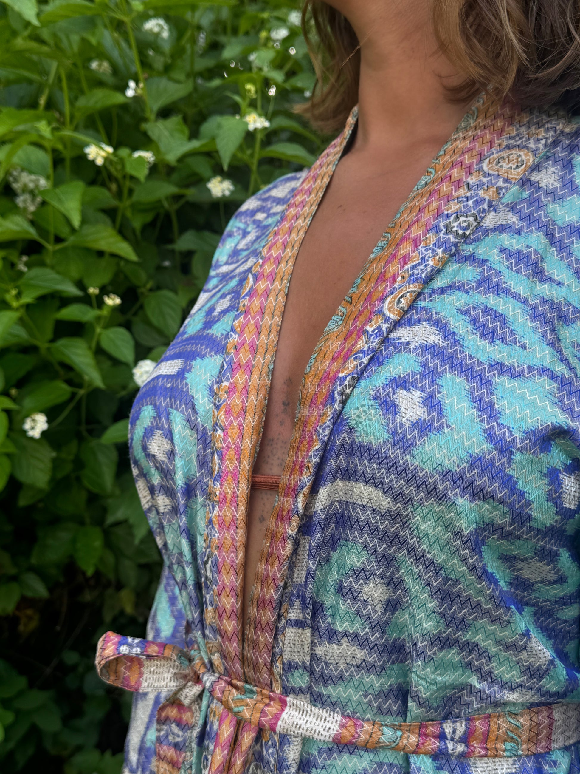 A close-up of a woman wearing a colorful patterned wrap robe with a tie waist, set against a background of green foliage and small white flowers.