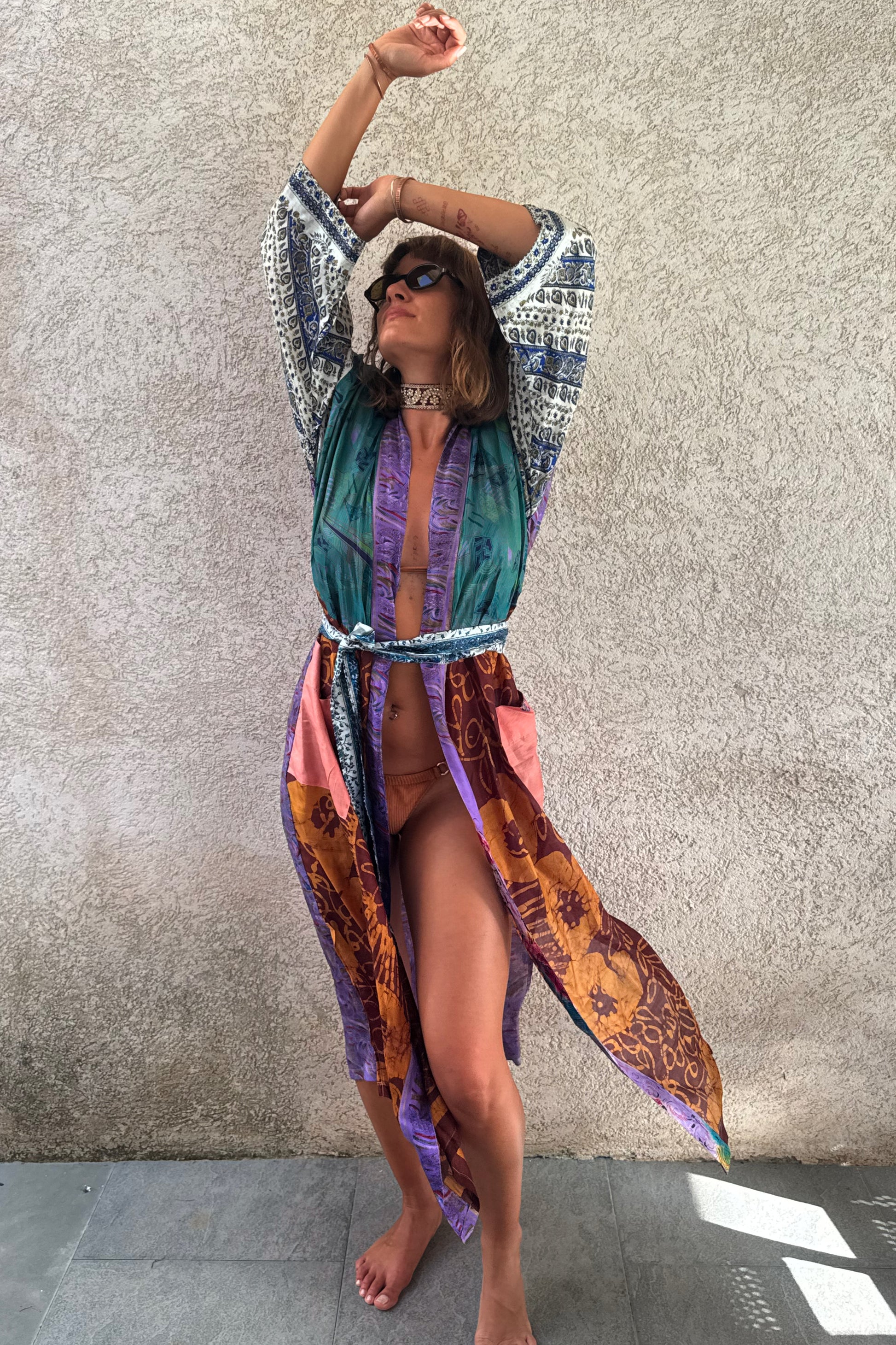 A woman modeling a colorful bohemian-style sheer maxi cover-up robe with a teal green, purple, pink, and orange paisley print, standing against a textured concrete wall.