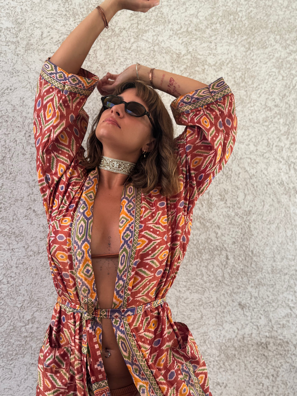 A woman wearing a bohemian-style patterned robe with orange, red, yellow, and blue geometric designs, styled with a silver choker and sunglasses, posing against a textured light gray wall.
