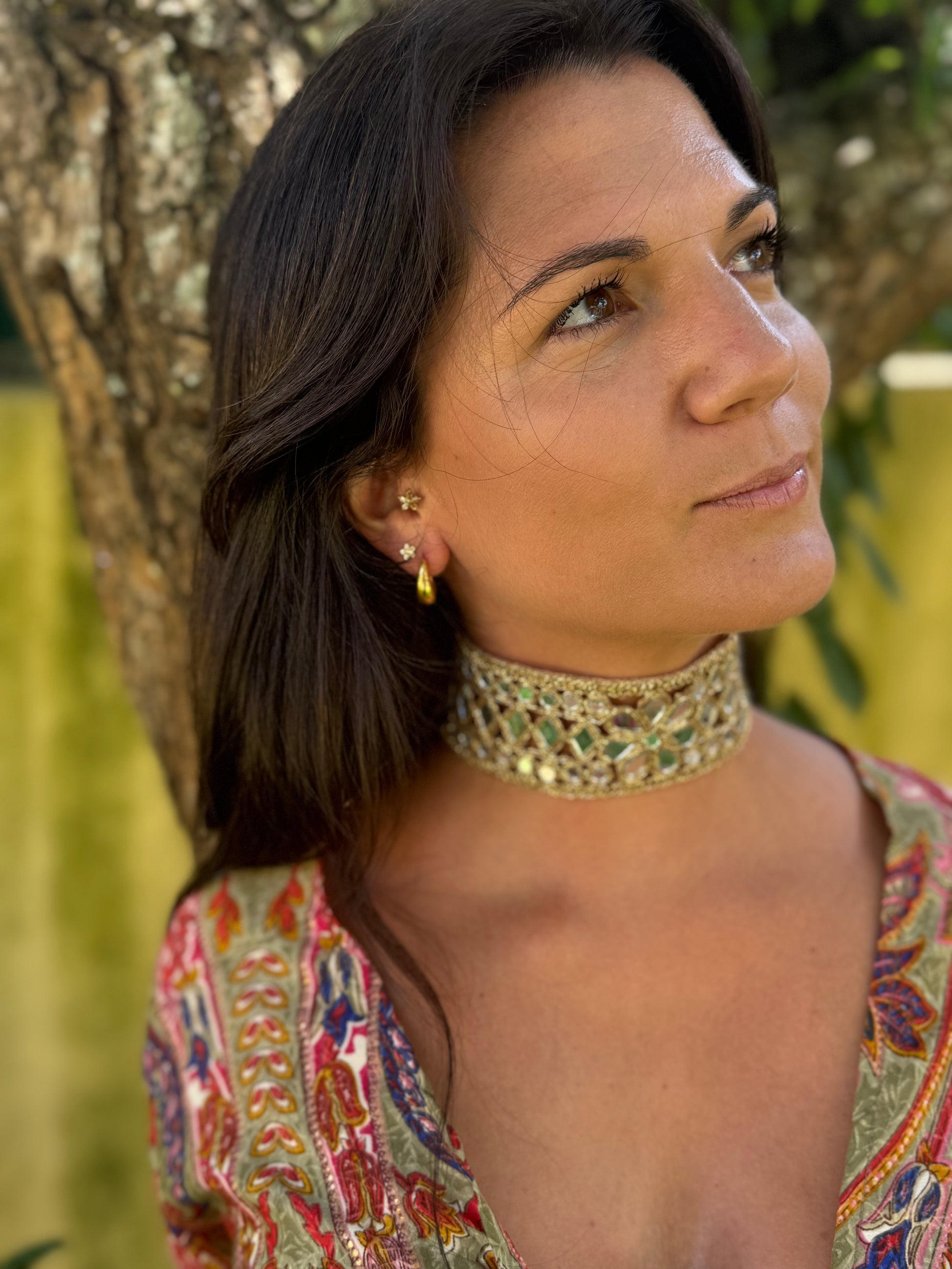 Woman wearing a colorful patterned top and gold choker necklace, standing against a blurred natural background.