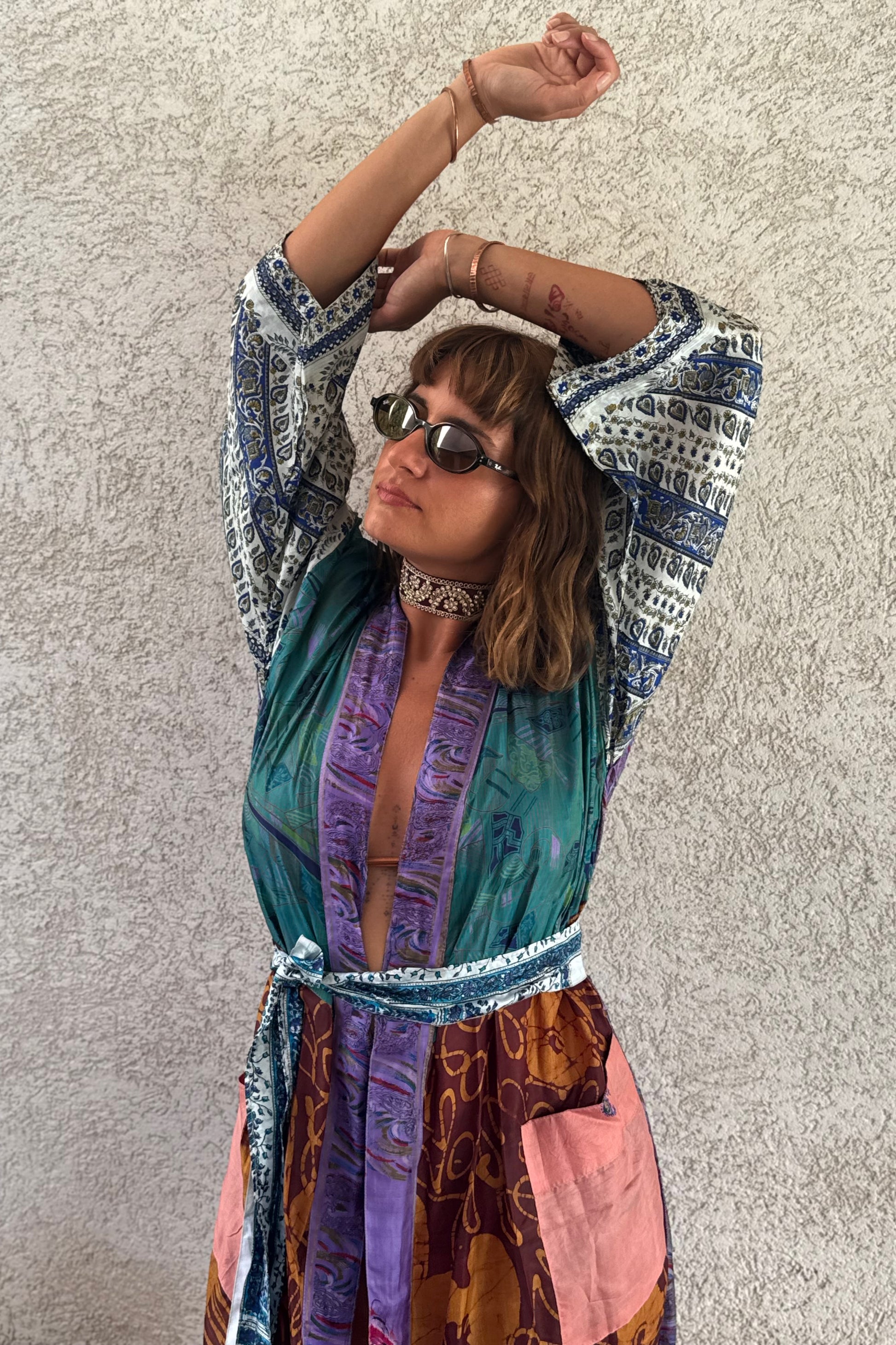 A woman wearing a colorful bohemian-style printed maxi dress with a tie waist and long sleeves, accessorized with a choker necklace and sunglasses, posing against a textured gray wall.
