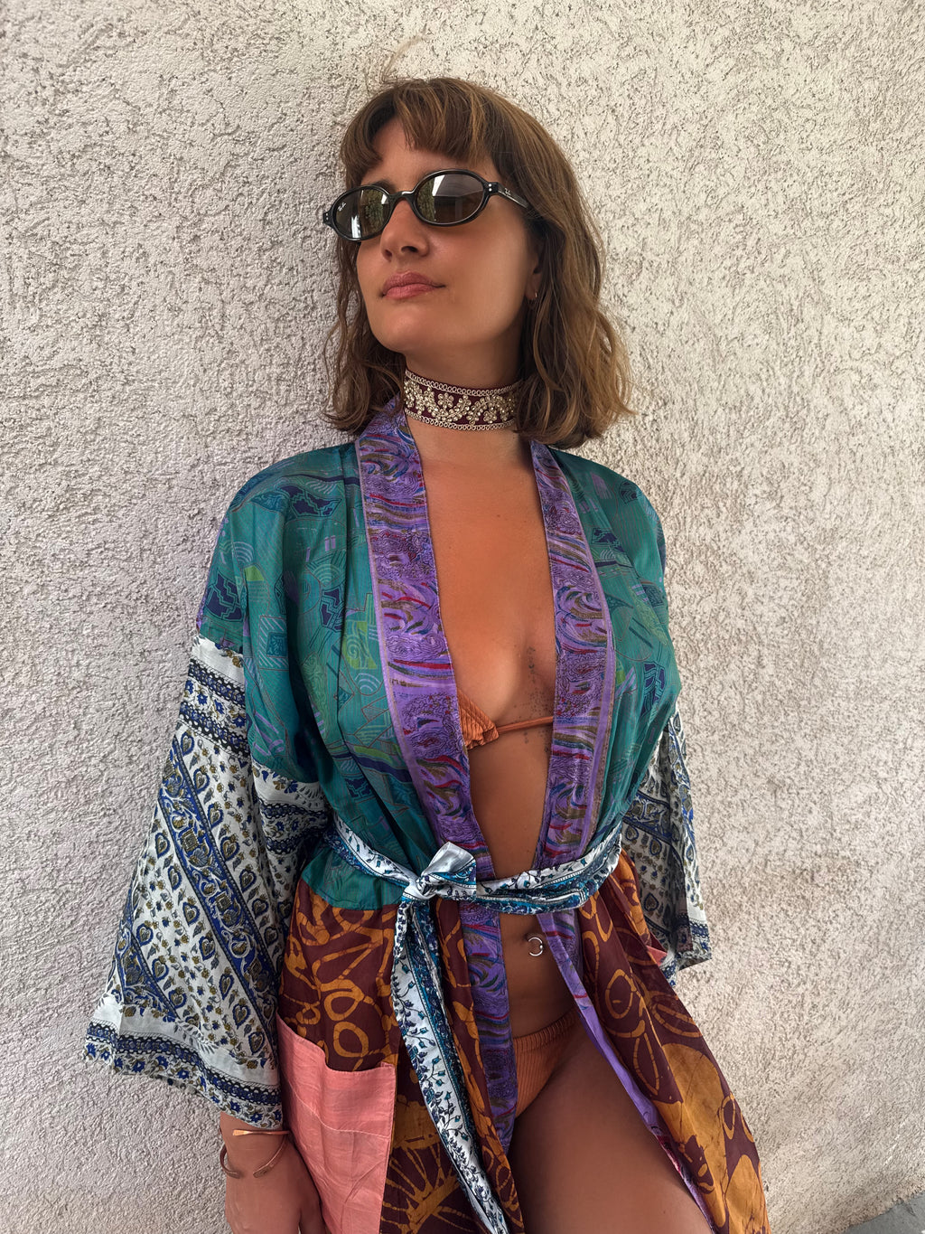 A woman wearing a colorful patterned open-front kimono-style cover-up with blue, purple, teal, white, and brown motifs, paired with sunglasses and a choker, posing against a textured light gray wall.