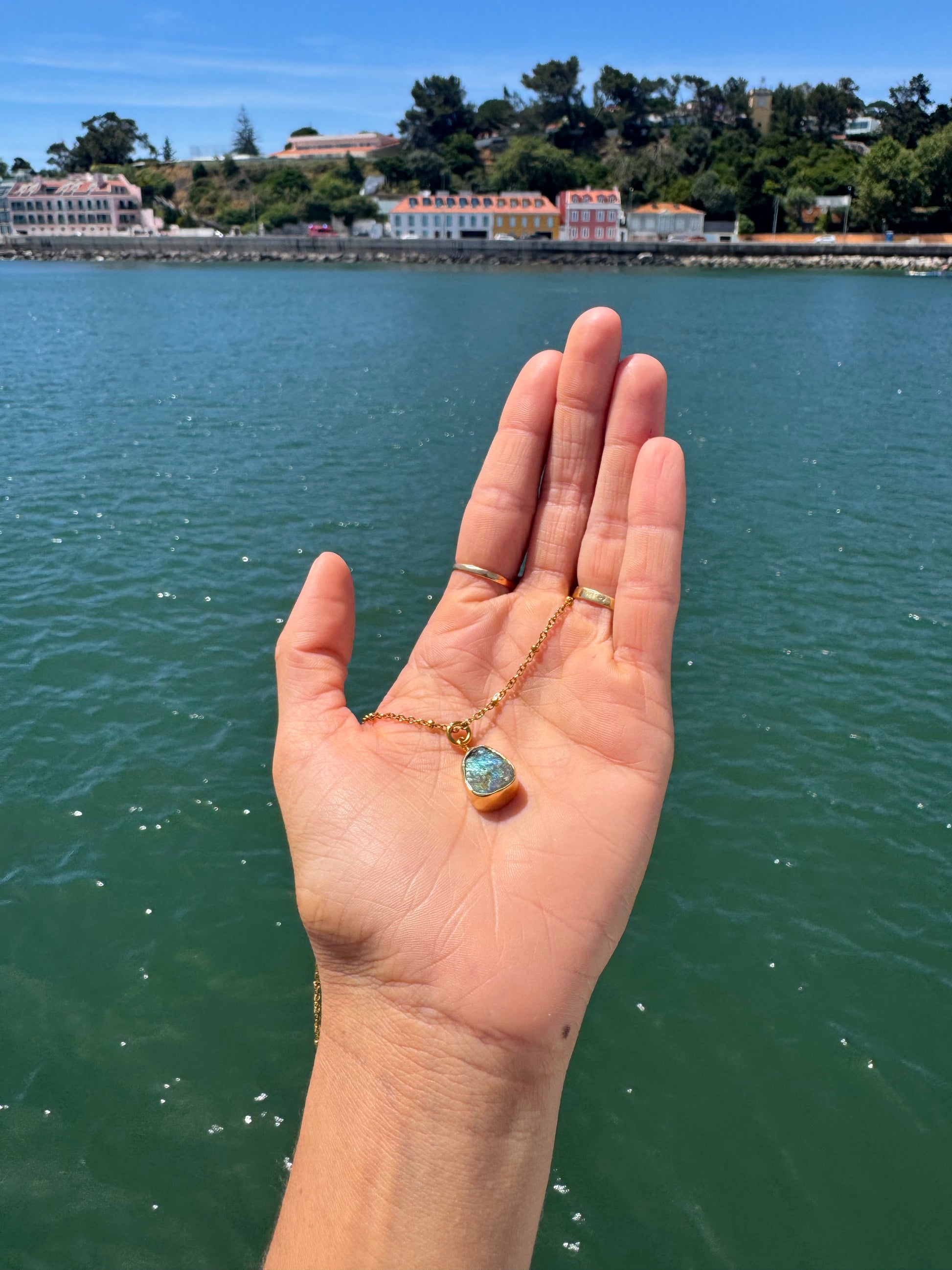 Hand holding a necklace with a blue pendant against a scenic waterfront background