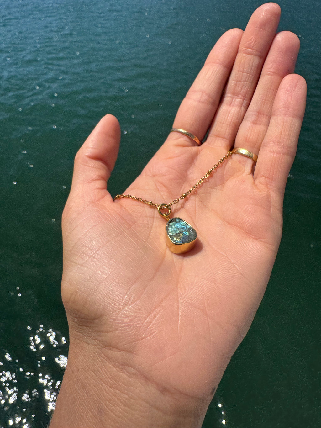 Hand holding a gold necklace with a blue pendant against a water background