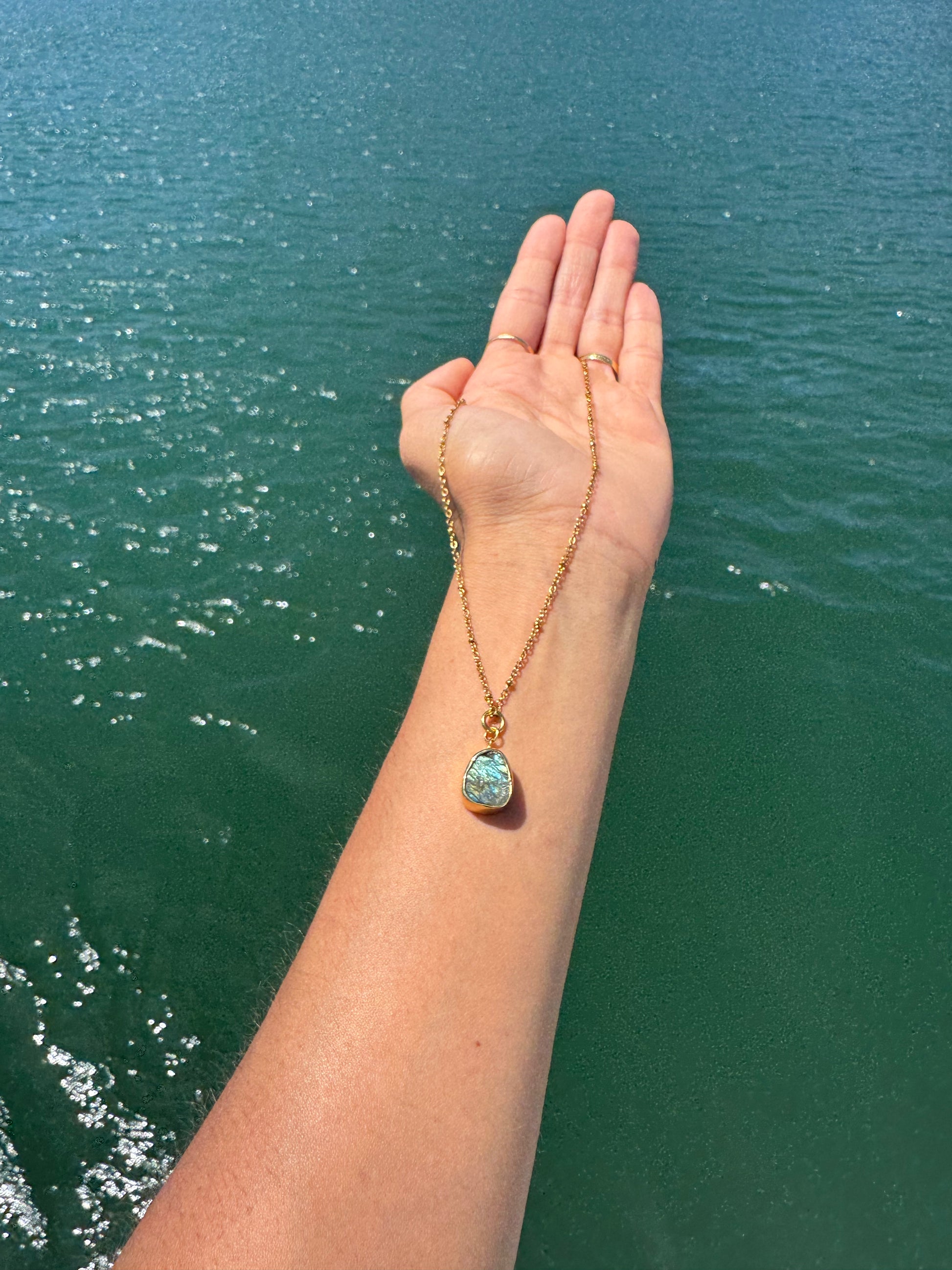 Hand wearing a gold necklace with a blue pendant against a body of water