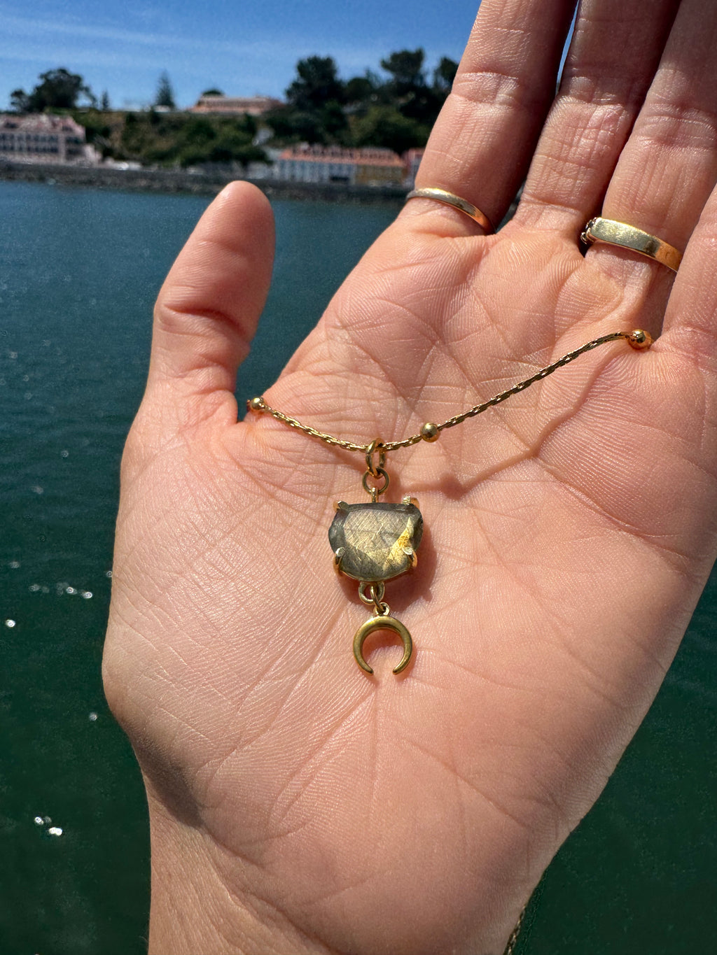Hand holding a gold necklace with a green stone pendant against a scenic background.