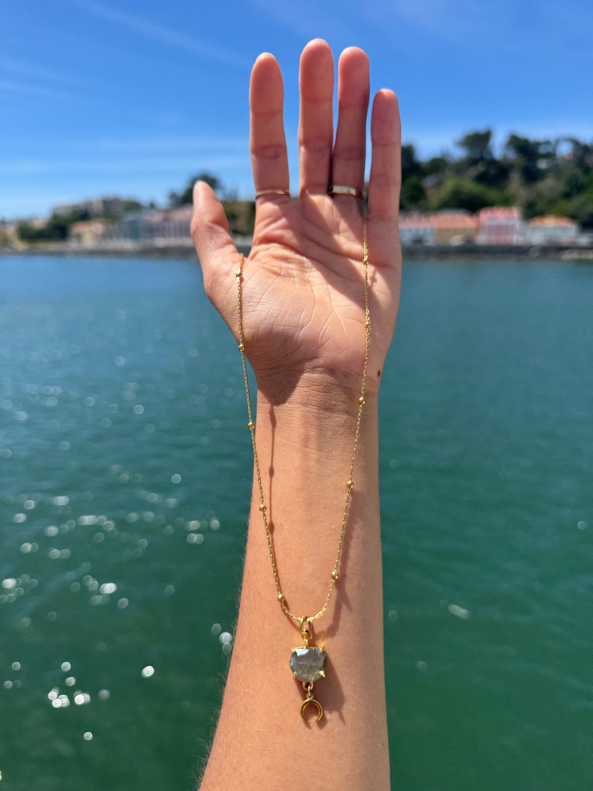 Hand wearing a gold necklace with a heart pendant against a scenic background