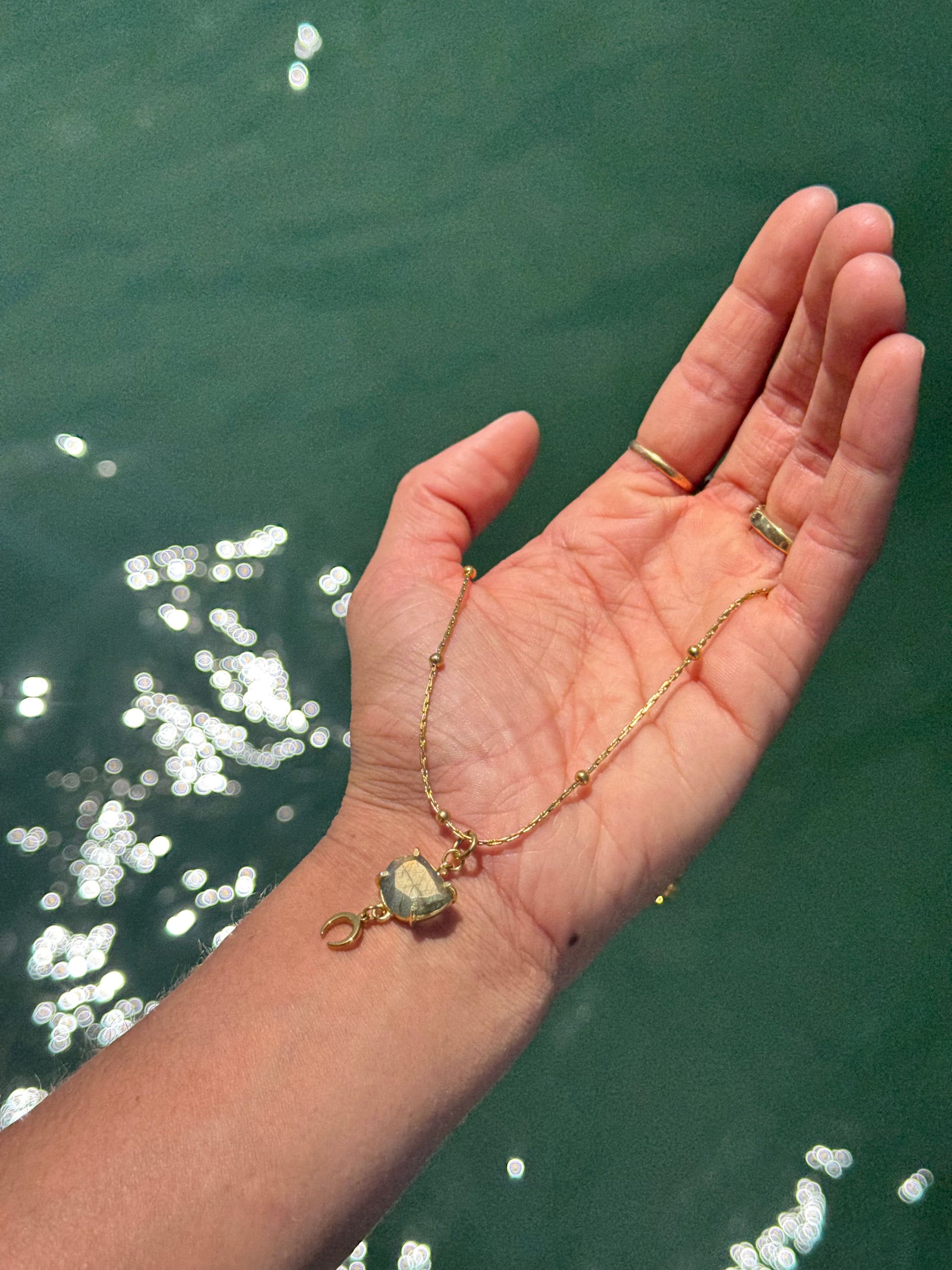 Hand holding a gold necklace with a pendant against a greenish water background