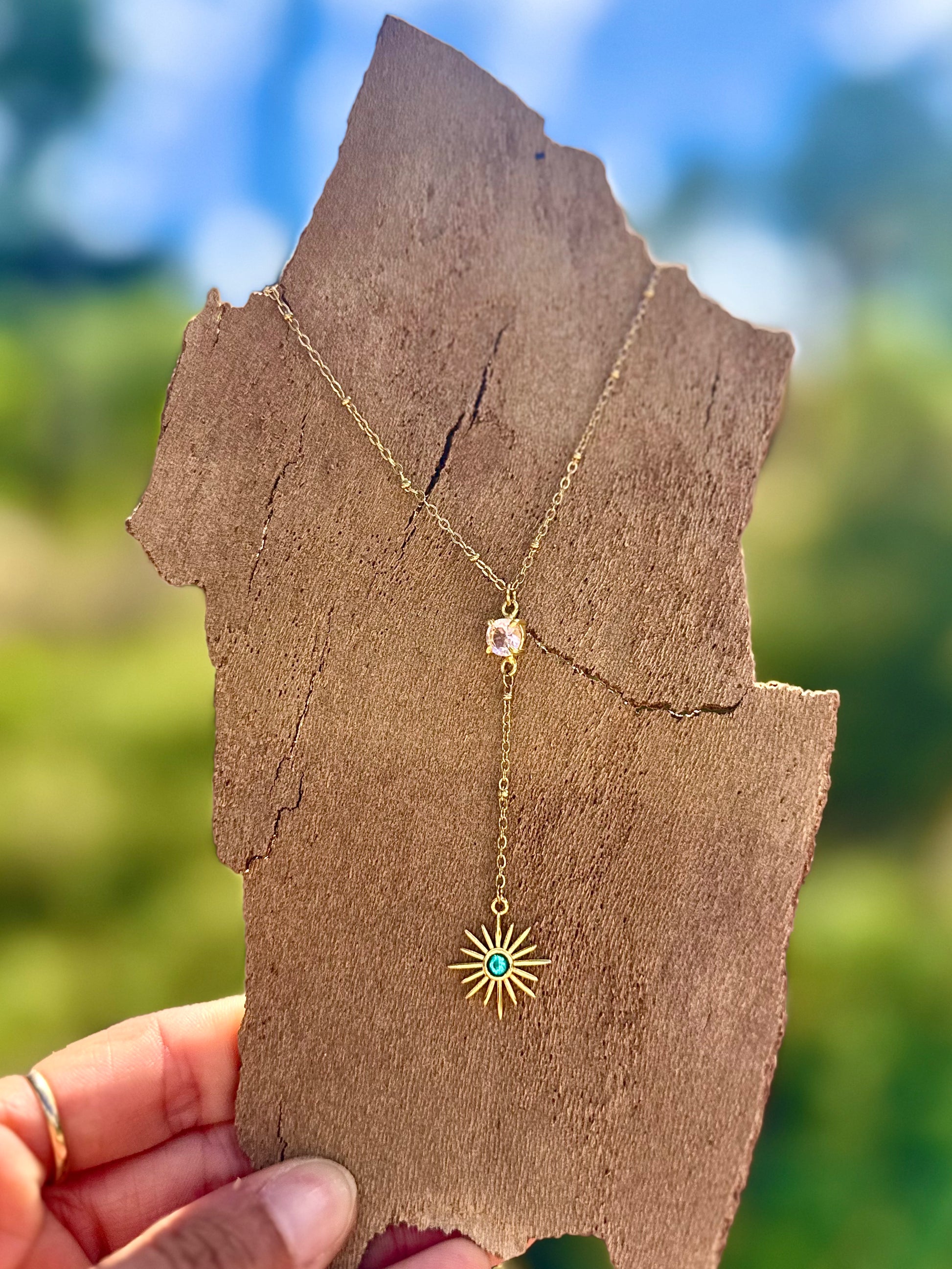 Gold necklace with a sunflower pendant held against a blurred natural background