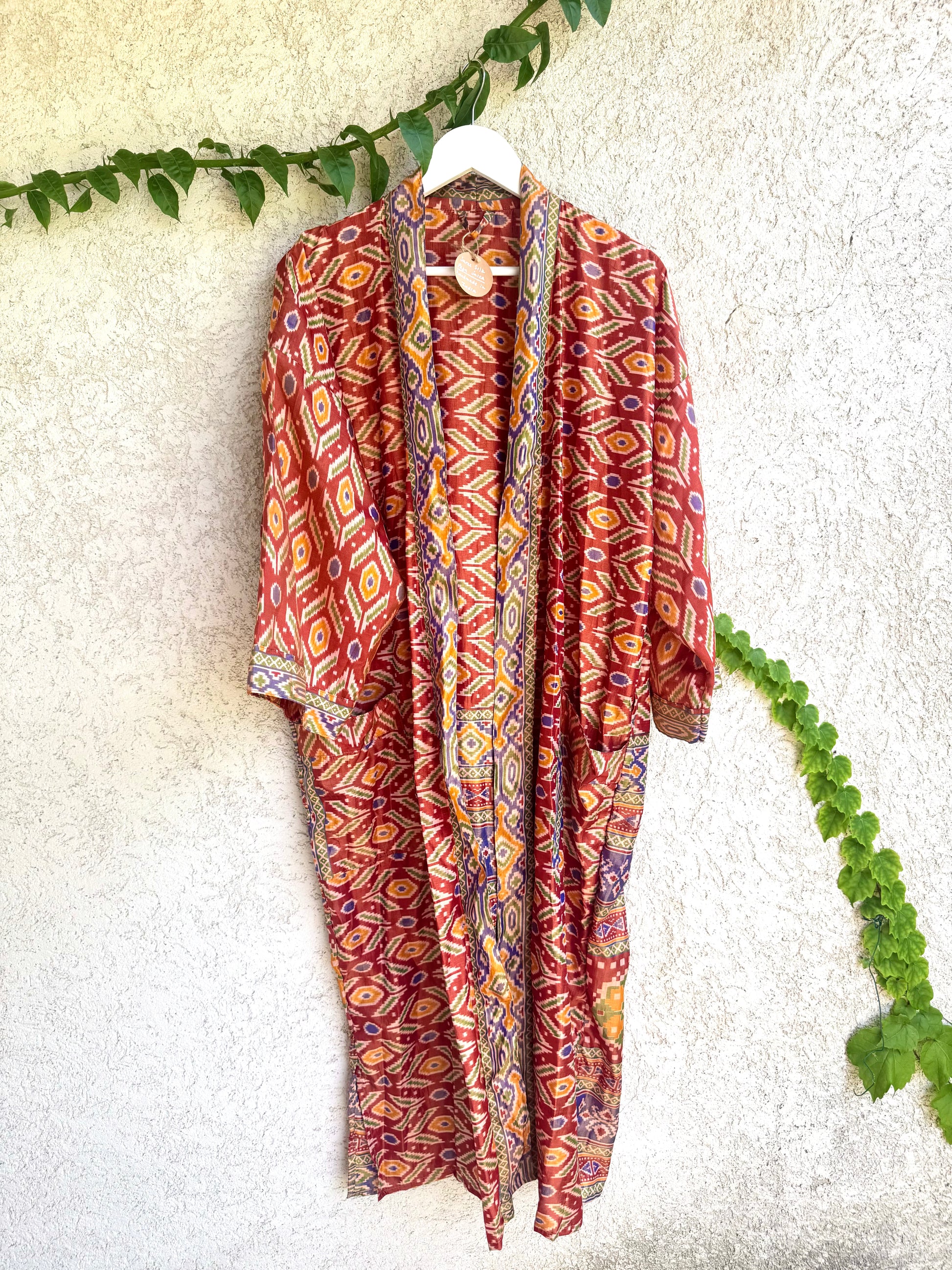 A lightweight open-front kimono-style cover-up with a vibrant red, orange, yellow, and blue geometric pattern, hanging on a white hanger against a textured off-white wall with green ivy vines.