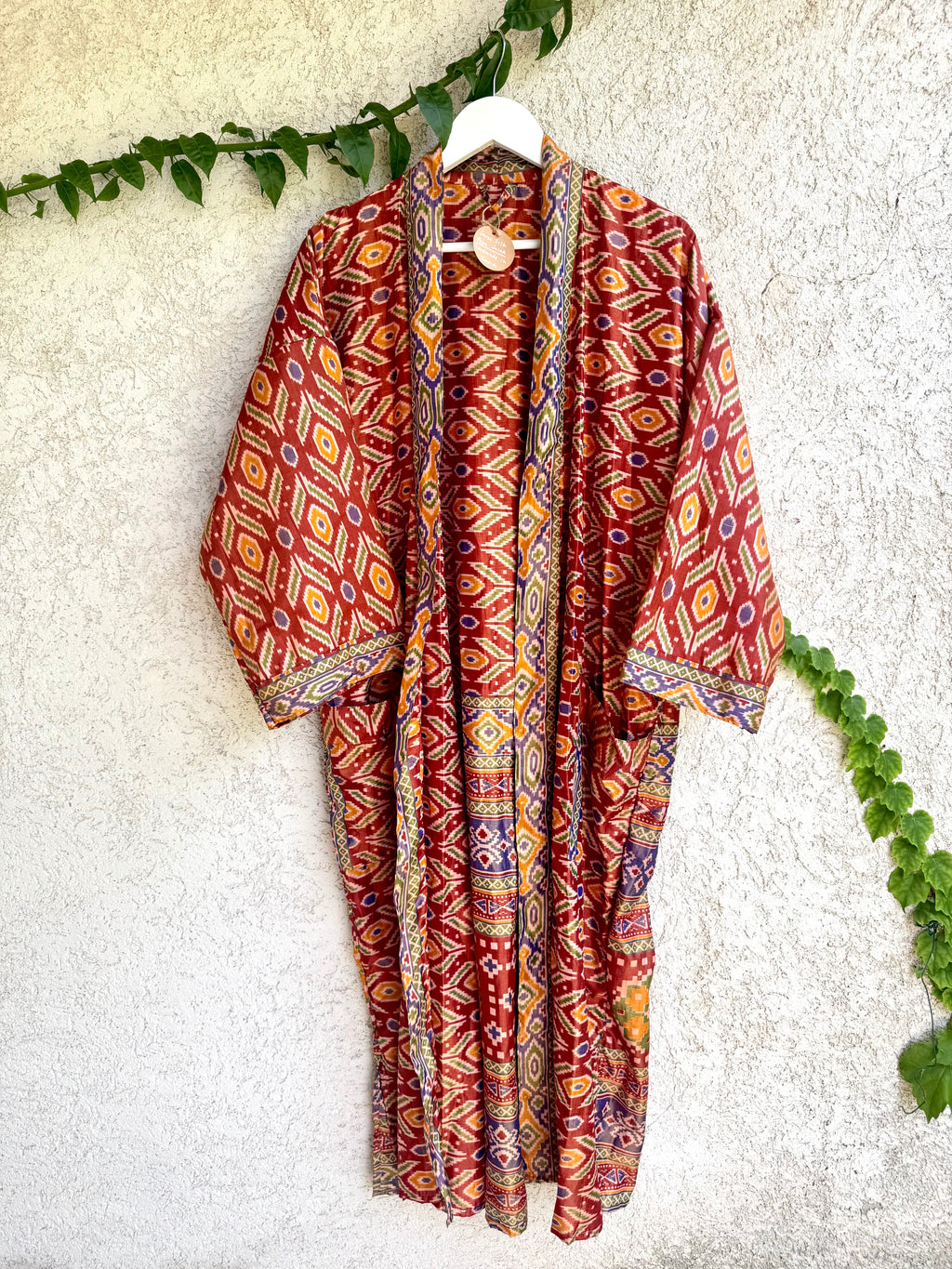 A lightweight open-front kimono robe with a vibrant red, orange, and blue geometric diamond pattern, hanging on a white hanger against a textured white stucco wall with green ivy vines.