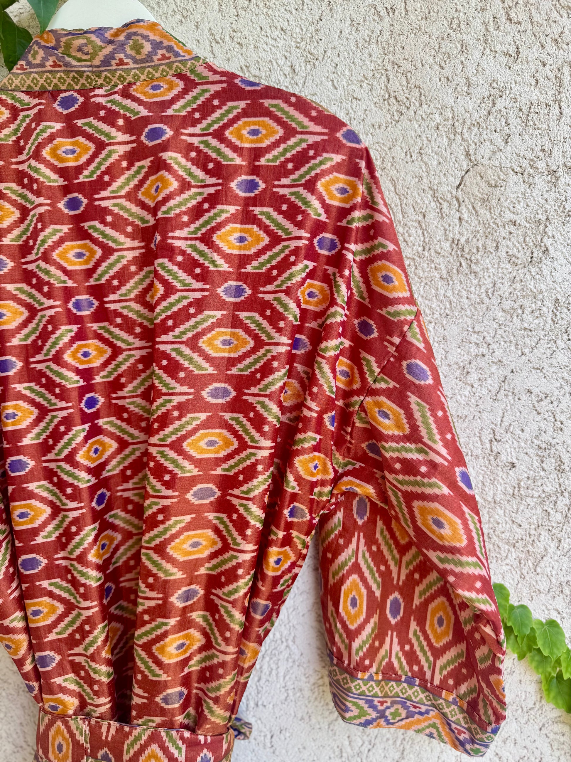 A long-sleeve collared shirt with a vibrant red base and a detailed geometric ikat pattern in orange, yellow, green, and purple, shown hanging against a textured beige stucco wall.