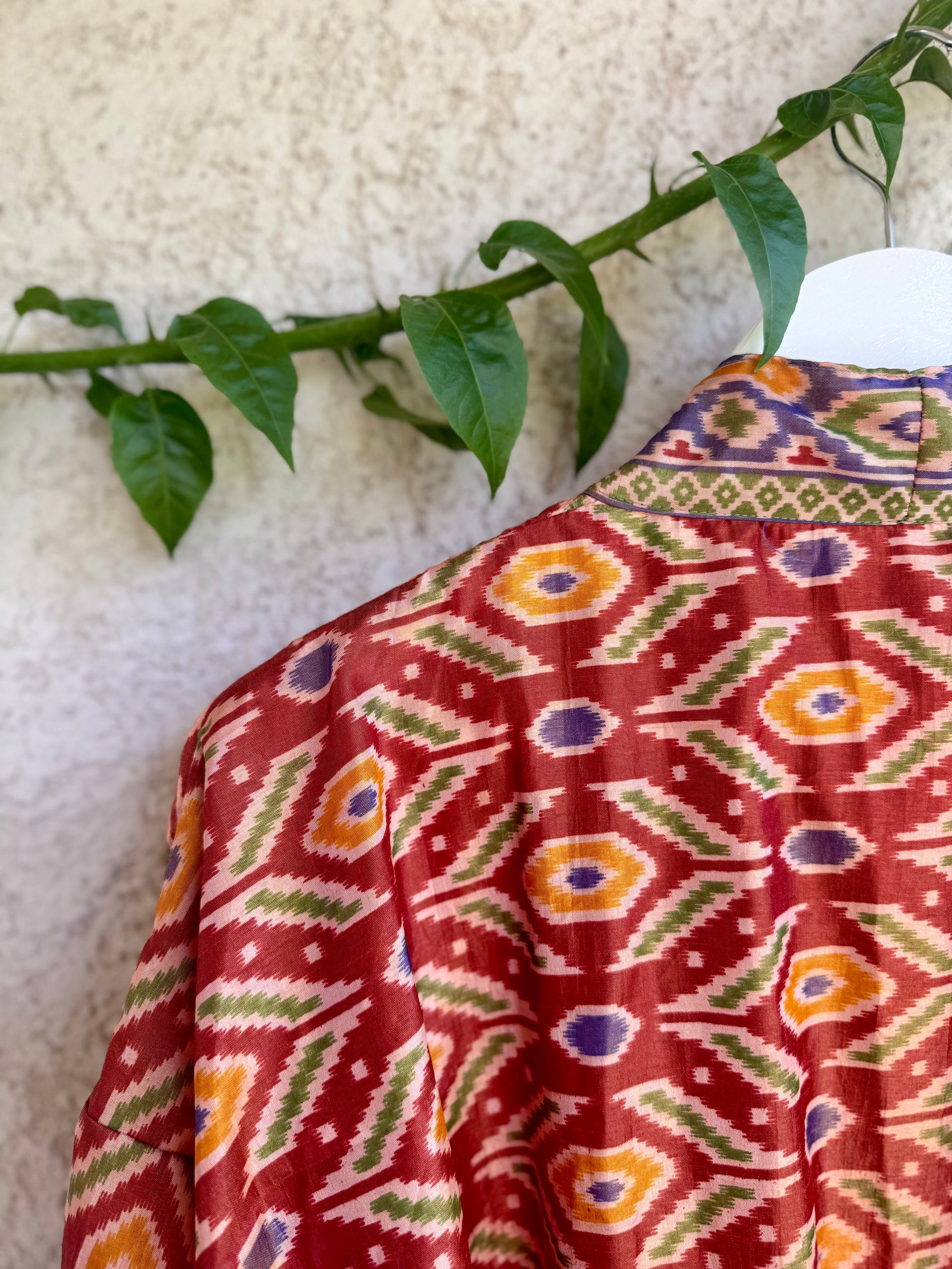 A close-up of a red patterned garment or fabric draped over a white hanger, featuring geometric ikat-style motifs in orange, green, purple, and cream, with a textured beige wall and green leaves in the background.