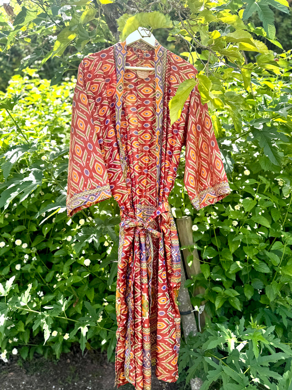 A red and orange patterned kimono robe with a matching tie belt, hanging on a white hanger outdoors against a background of green garden foliage.