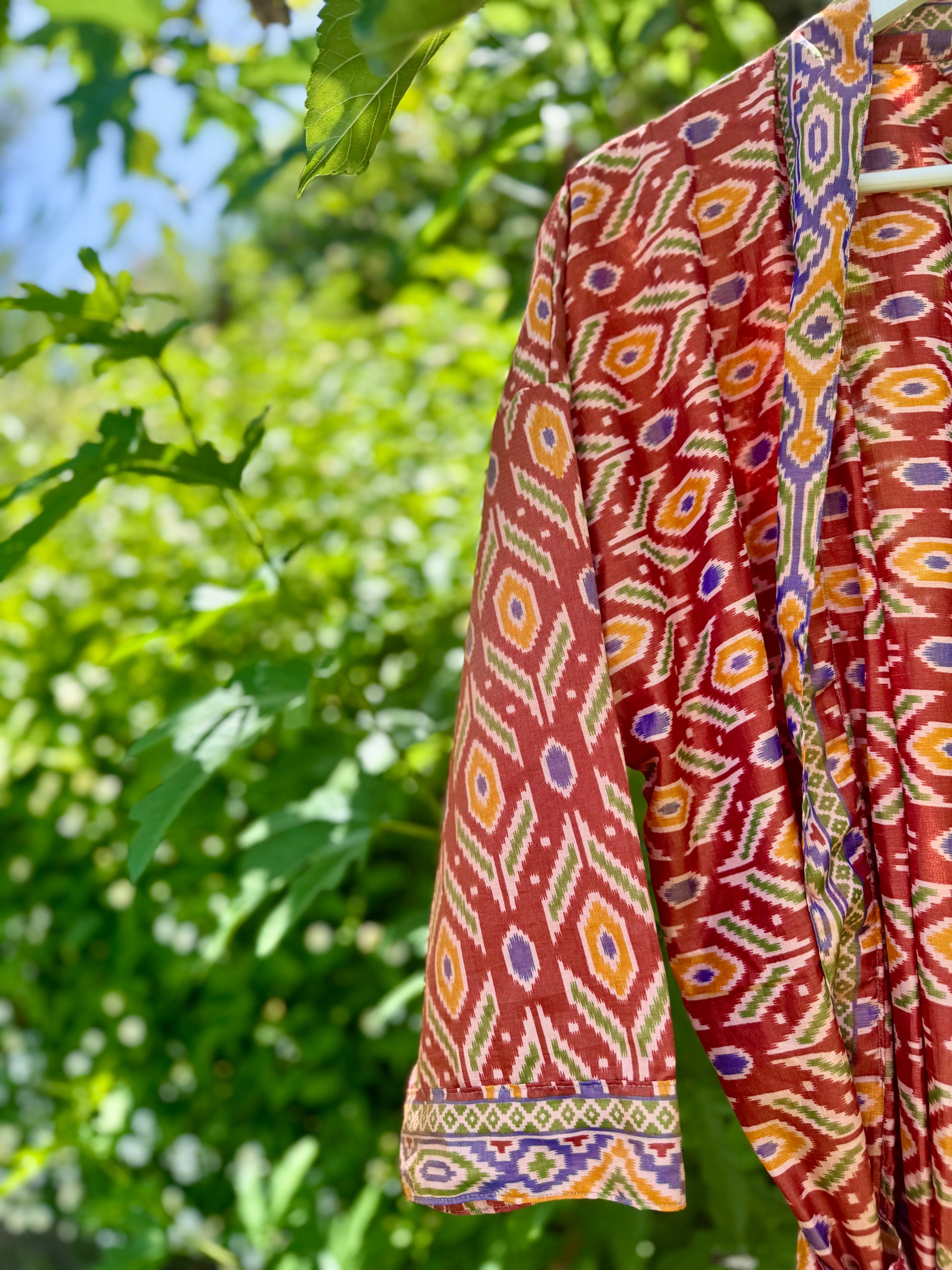 A close-up of a patterned garment hanging outdoors against a blurred background of green leaves and blue sky. The fabric features a geometric ikat-style design in rust red, orange, yellow, purple, and green.