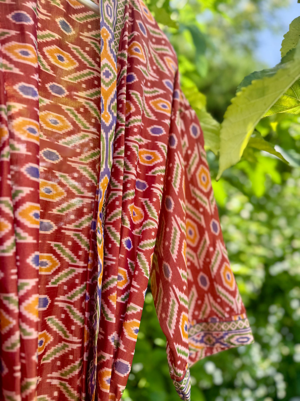 A close-up of a rust-red patterned garment with geometric ikat-style motifs in orange, yellow, green, and purple, shown outdoors against a blurred green leafy background.