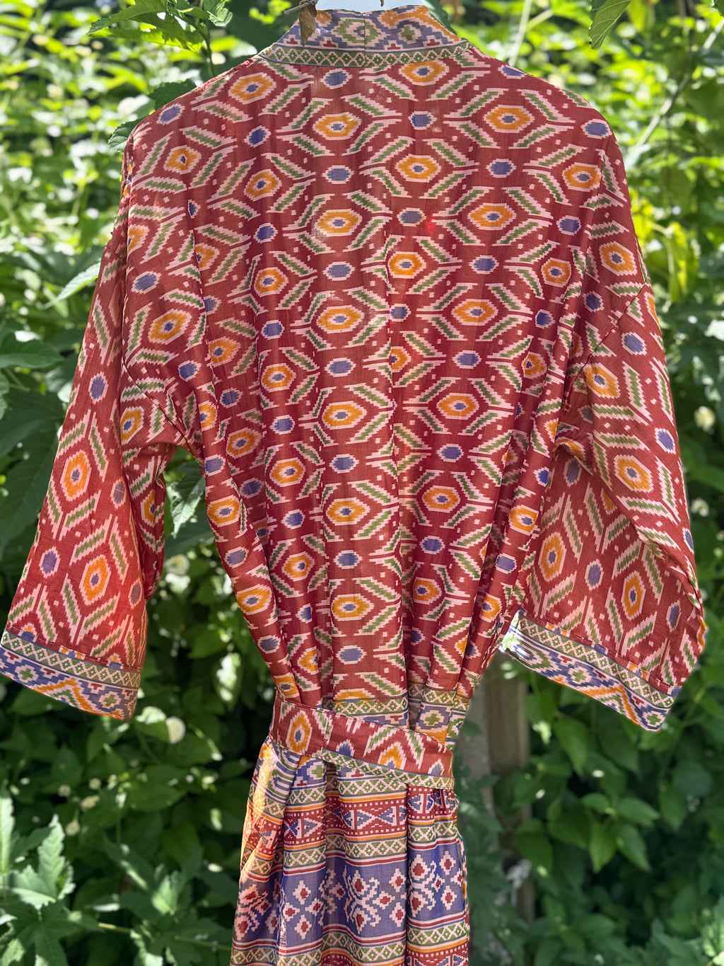 A red patterned kaftan dress with geometric motifs in orange, yellow, and blue, shown hanging outdoors against a background of green foliage.