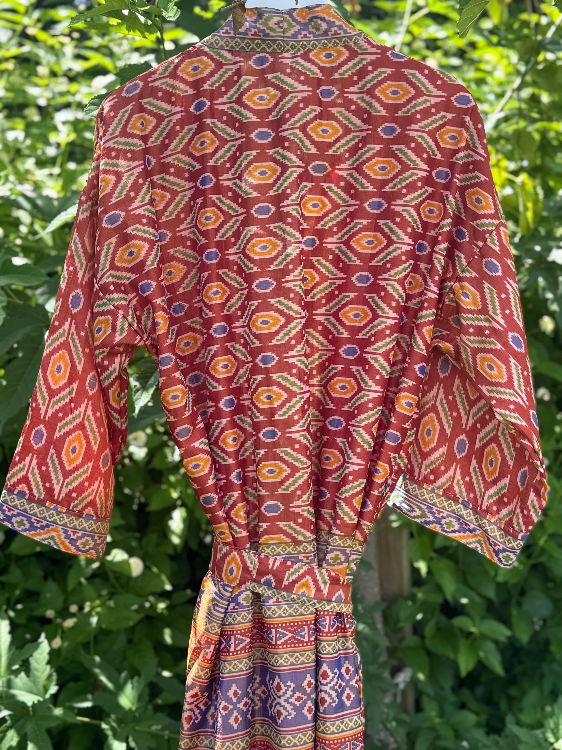 A red patterned kaftan dress with geometric motifs in orange, yellow, and blue, shown hanging outdoors against a background of green foliage.