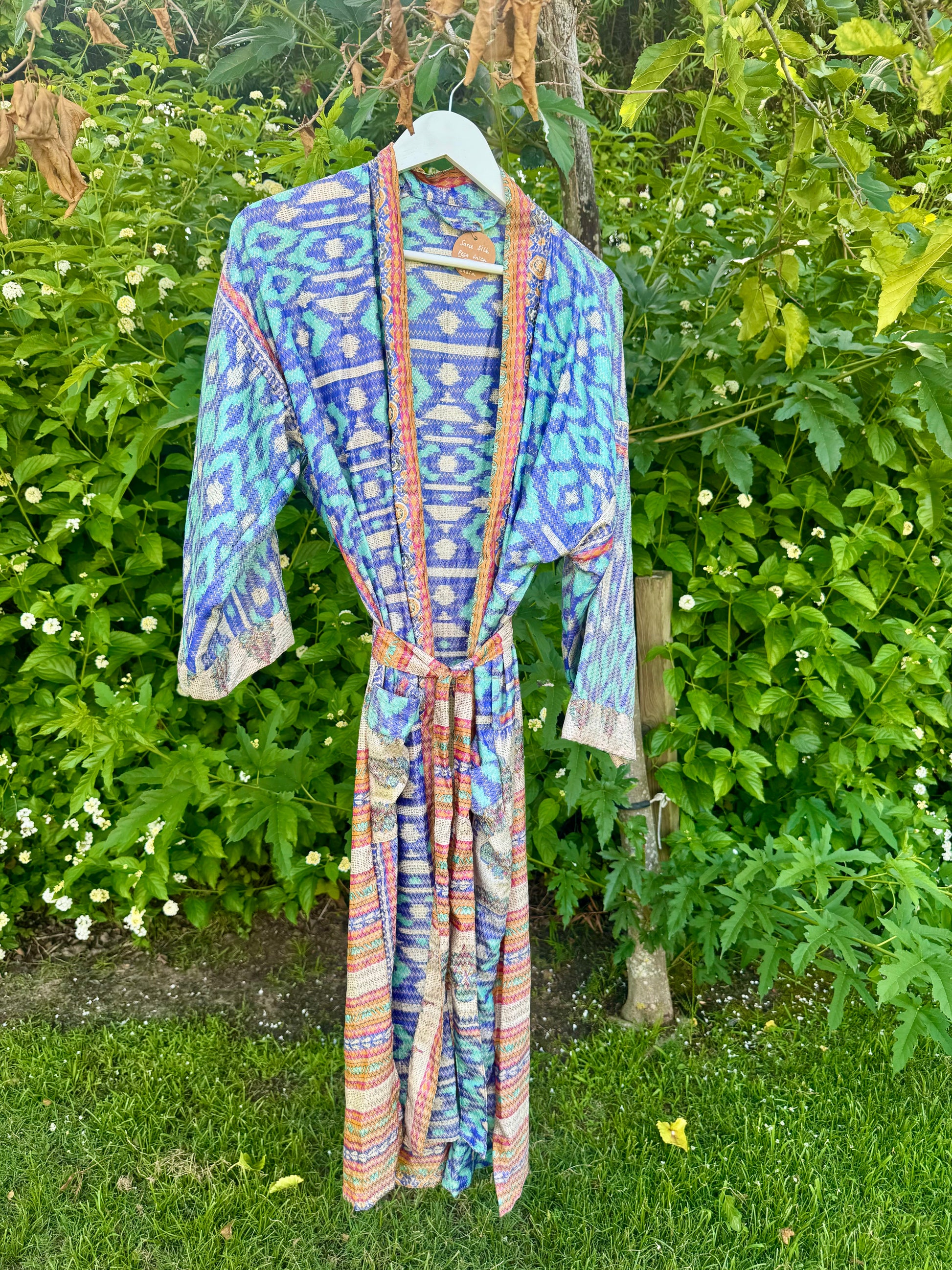 A long kaftan robe with a blue, purple, and orange geometric ikat pattern hangs on a white hanger outdoors against a backdrop of green garden foliage.