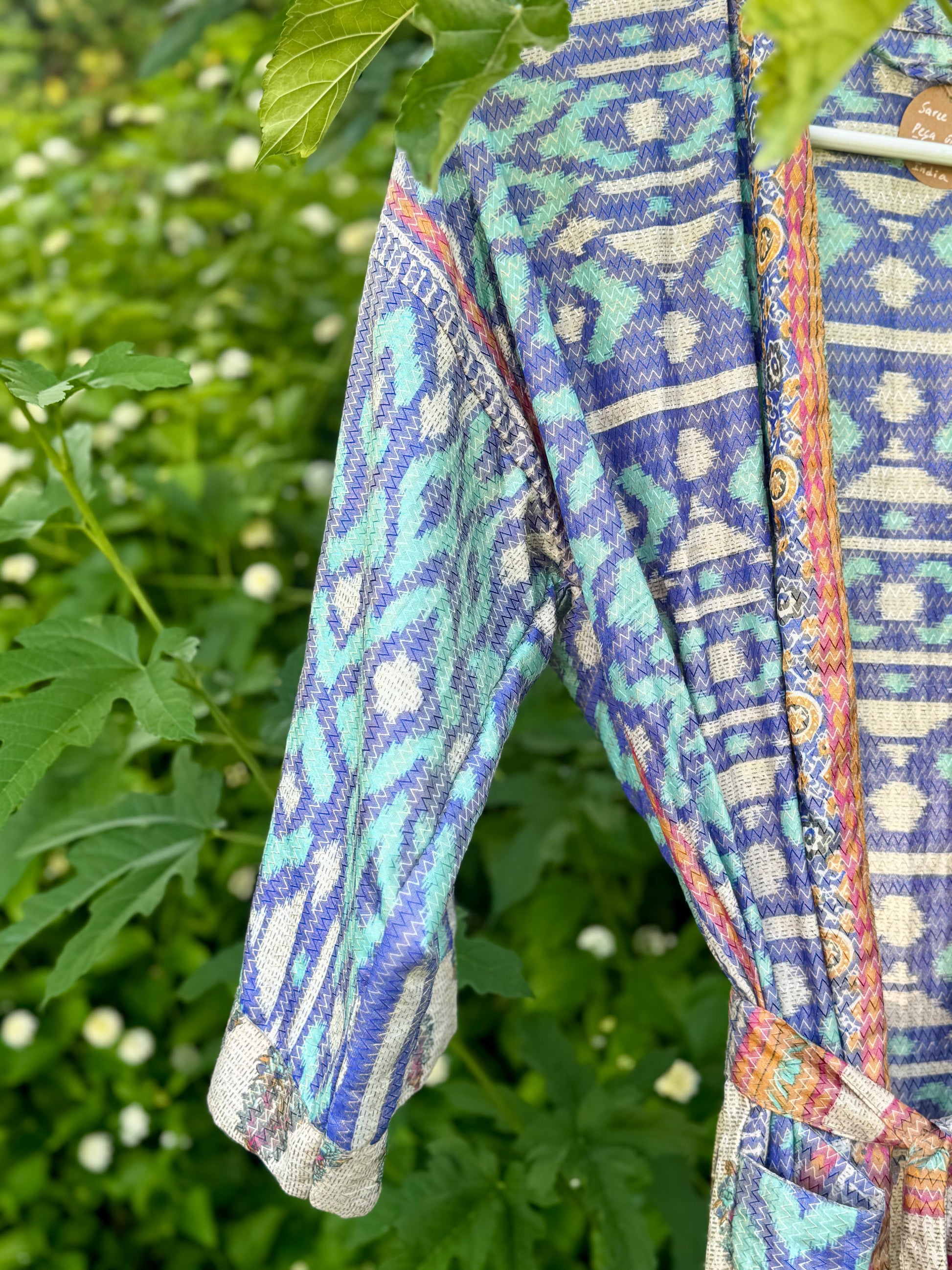 A patterned wrap robe with a blue, green, and white ikat design and a multicolor border, hanging outdoors against a background of green foliage.
