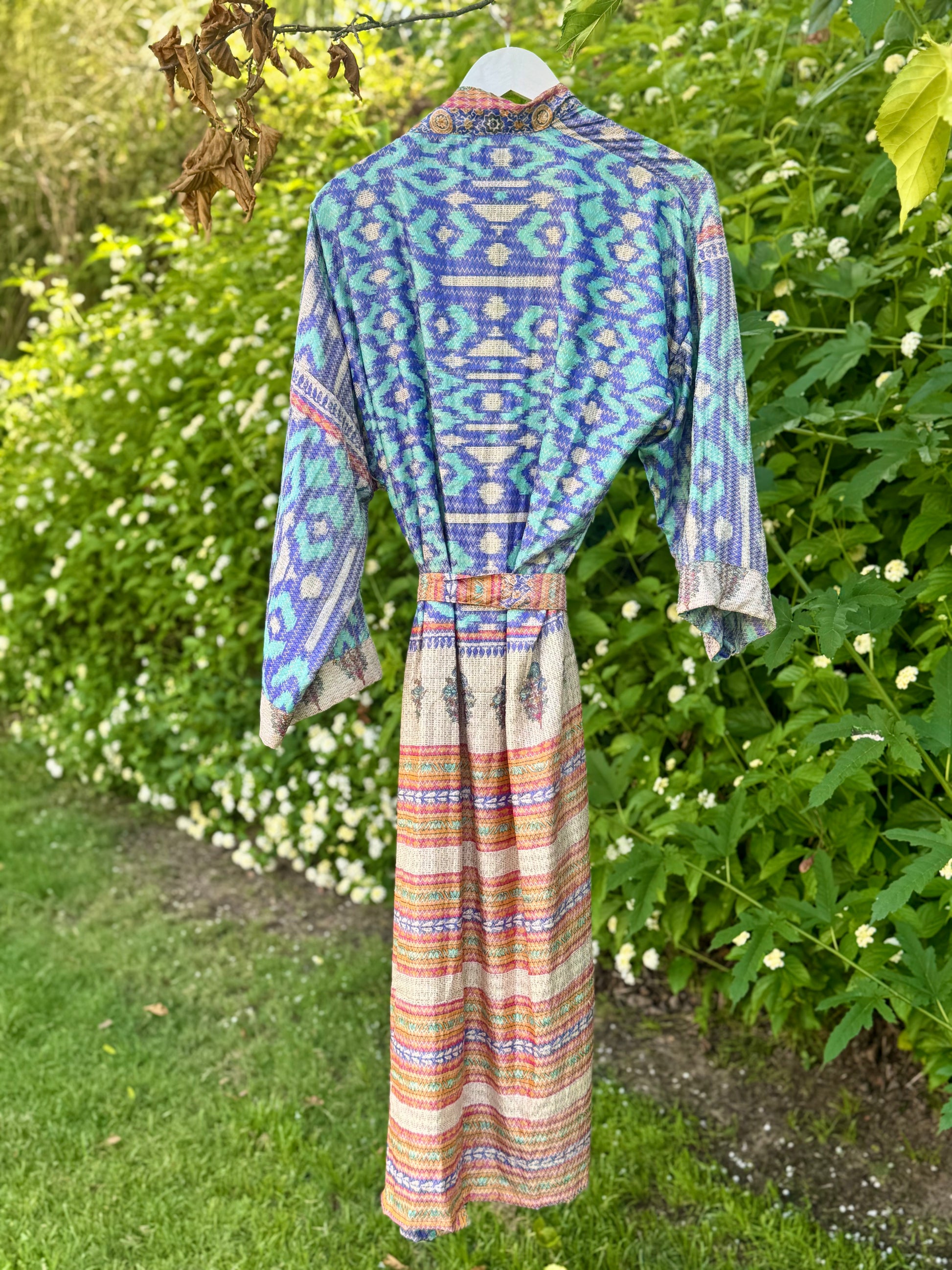 A long-sleeved maxi dress with a blue and purple geometric pattern on the bodice and multicolored striped skirt, hanging on a white hanger in a garden setting.