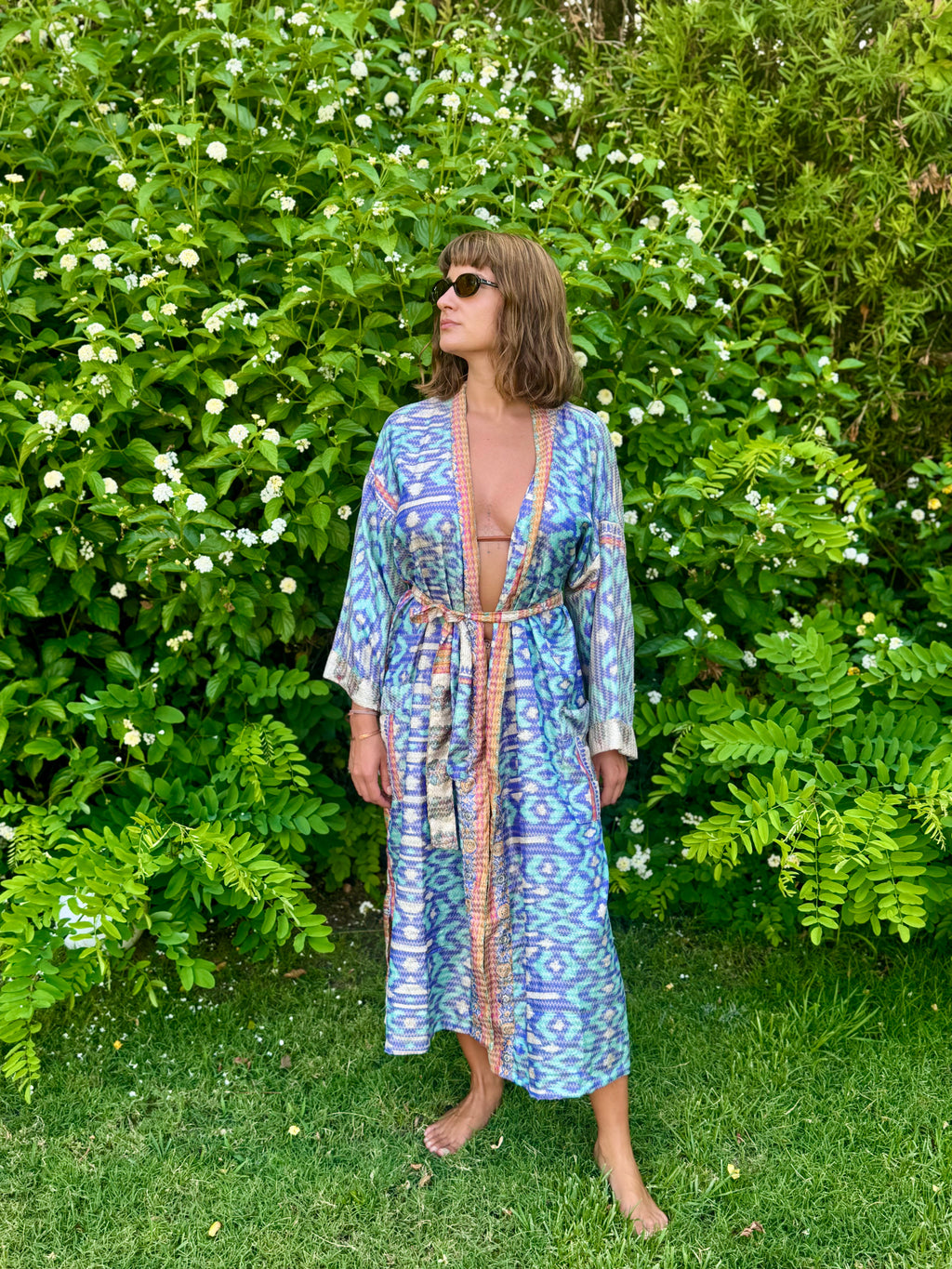 A woman wearing a blue and purple patterned kaftan robe with a beige border stands barefoot on a grassy lawn in front of a lush green garden wall with white flowers.