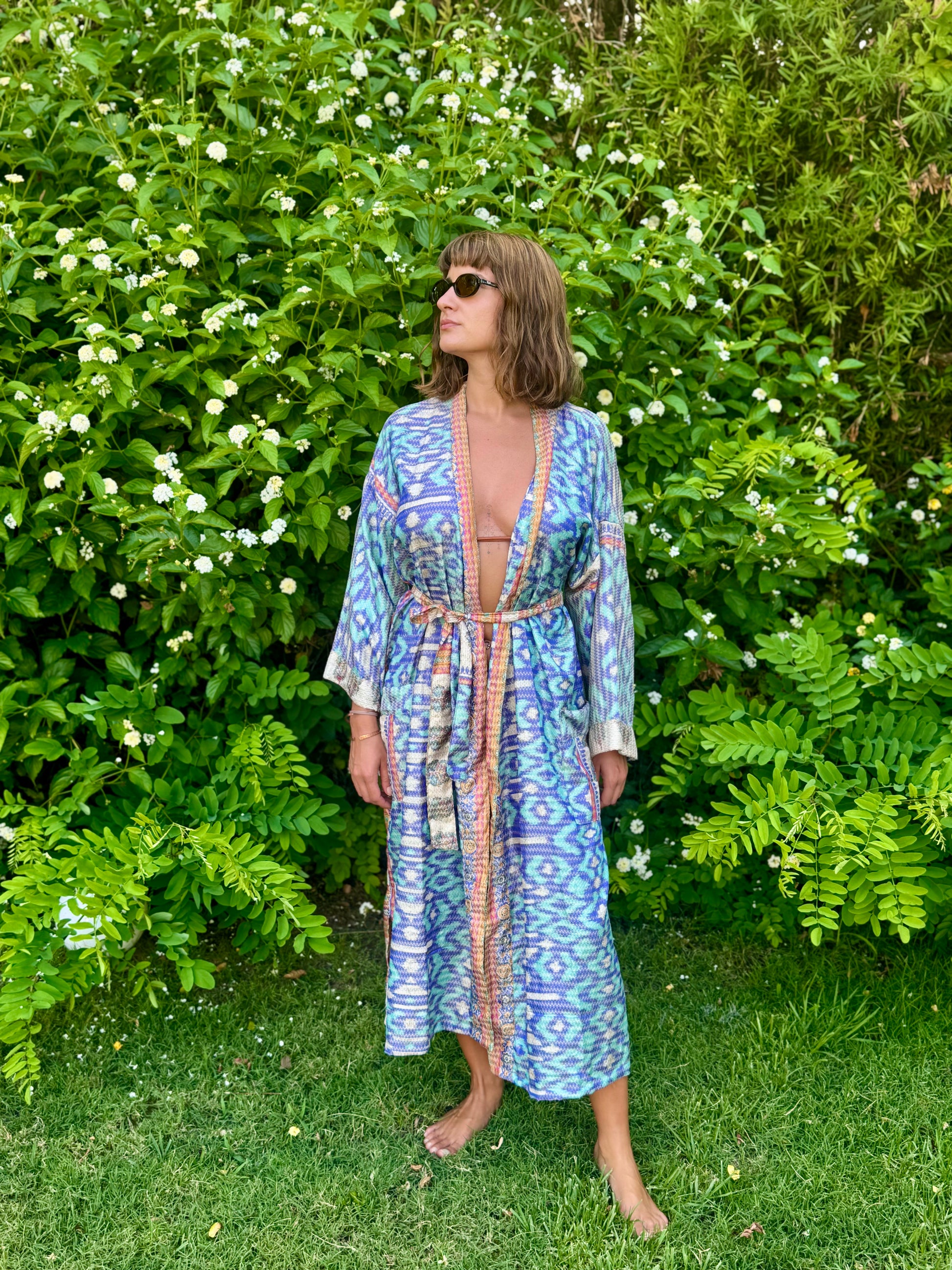 A woman wearing a blue and purple patterned kaftan robe with a beige border stands barefoot on a grassy lawn in front of a lush green garden wall with white flowers.