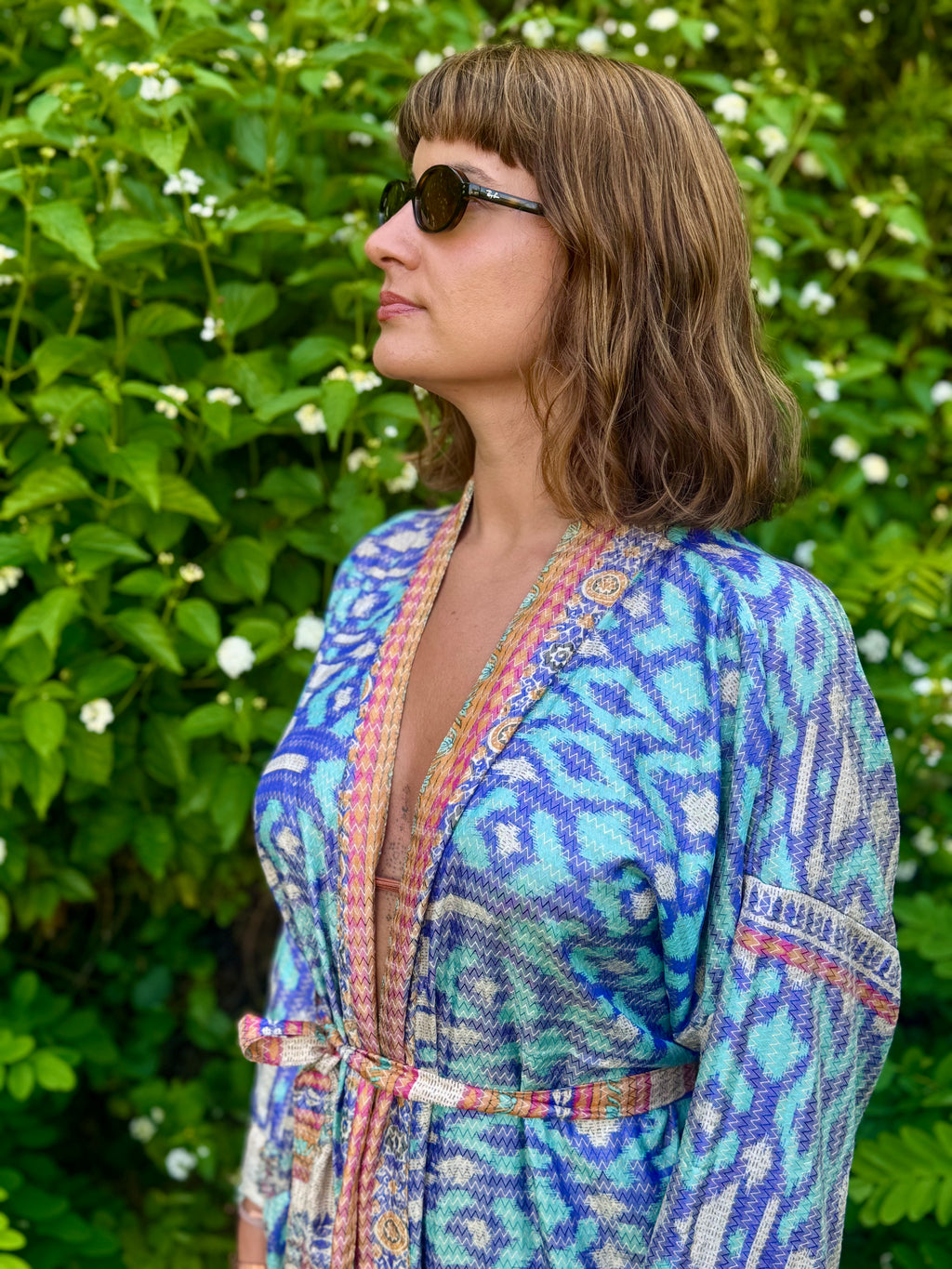 A woman wearing a blue and turquoise patterned wrap-style robe with multicolored trim, standing outdoors in front of a green leafy background.