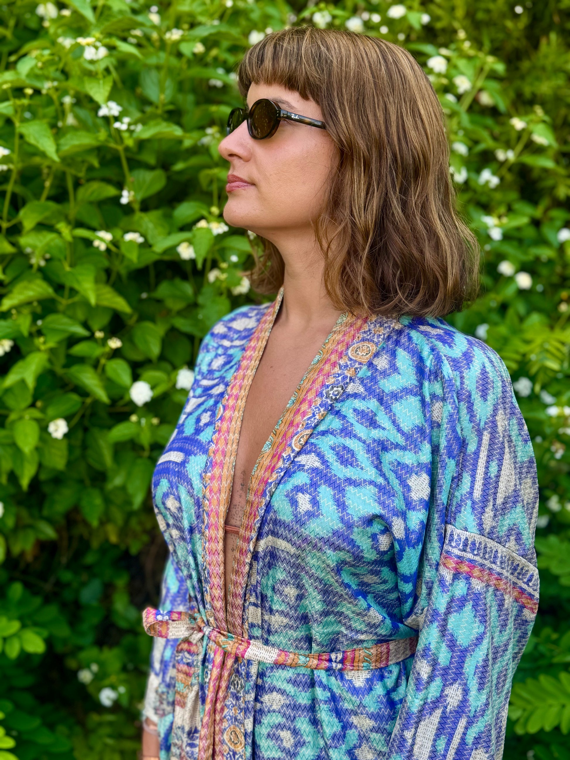 A woman wearing a blue and turquoise patterned wrap-style robe with multicolored trim, standing outdoors in front of a green leafy background.