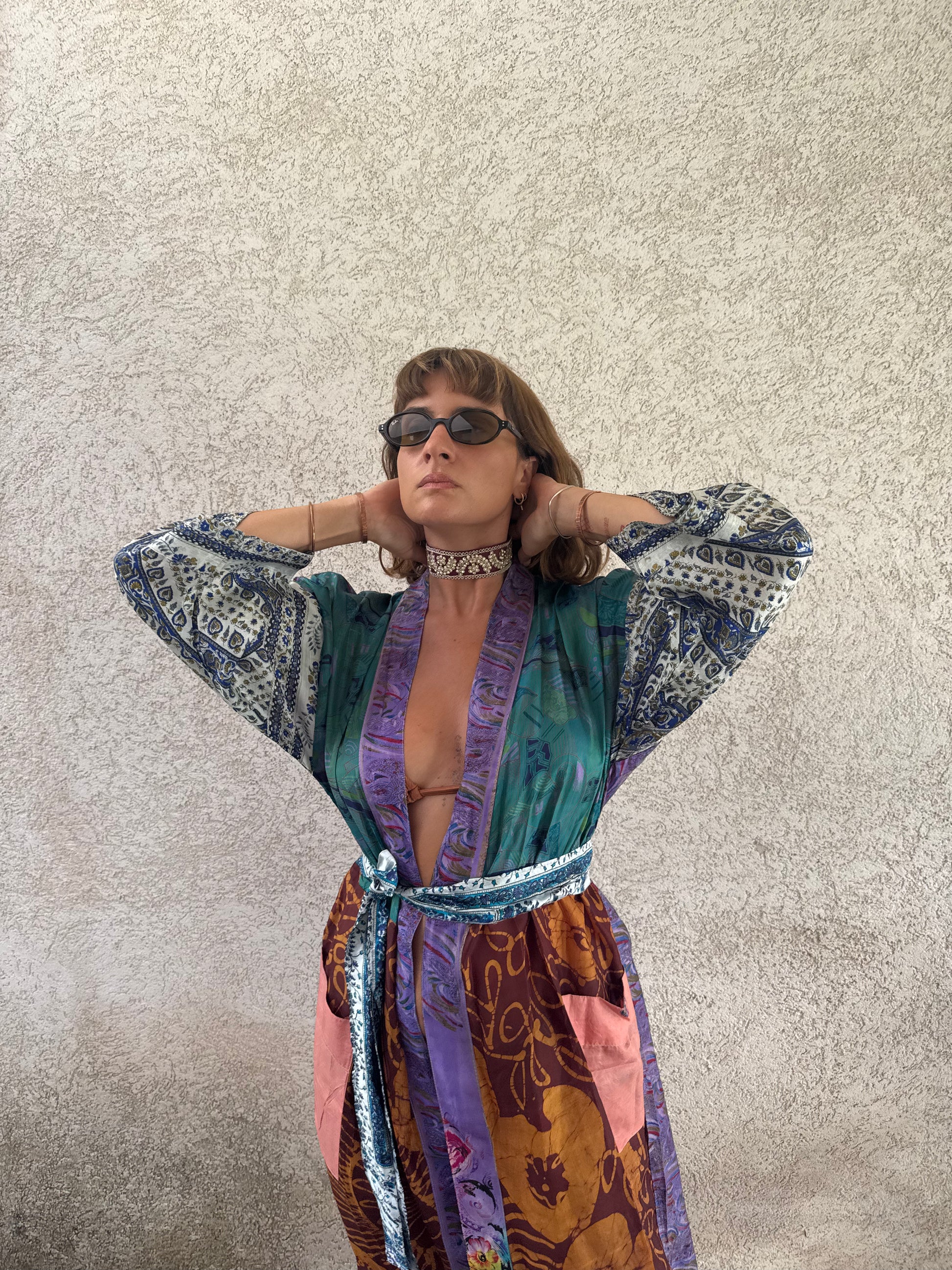 A person wearing a colorful patterned robe with wide sleeves, a tie belt, and a choker necklace, standing against a textured light gray wall.