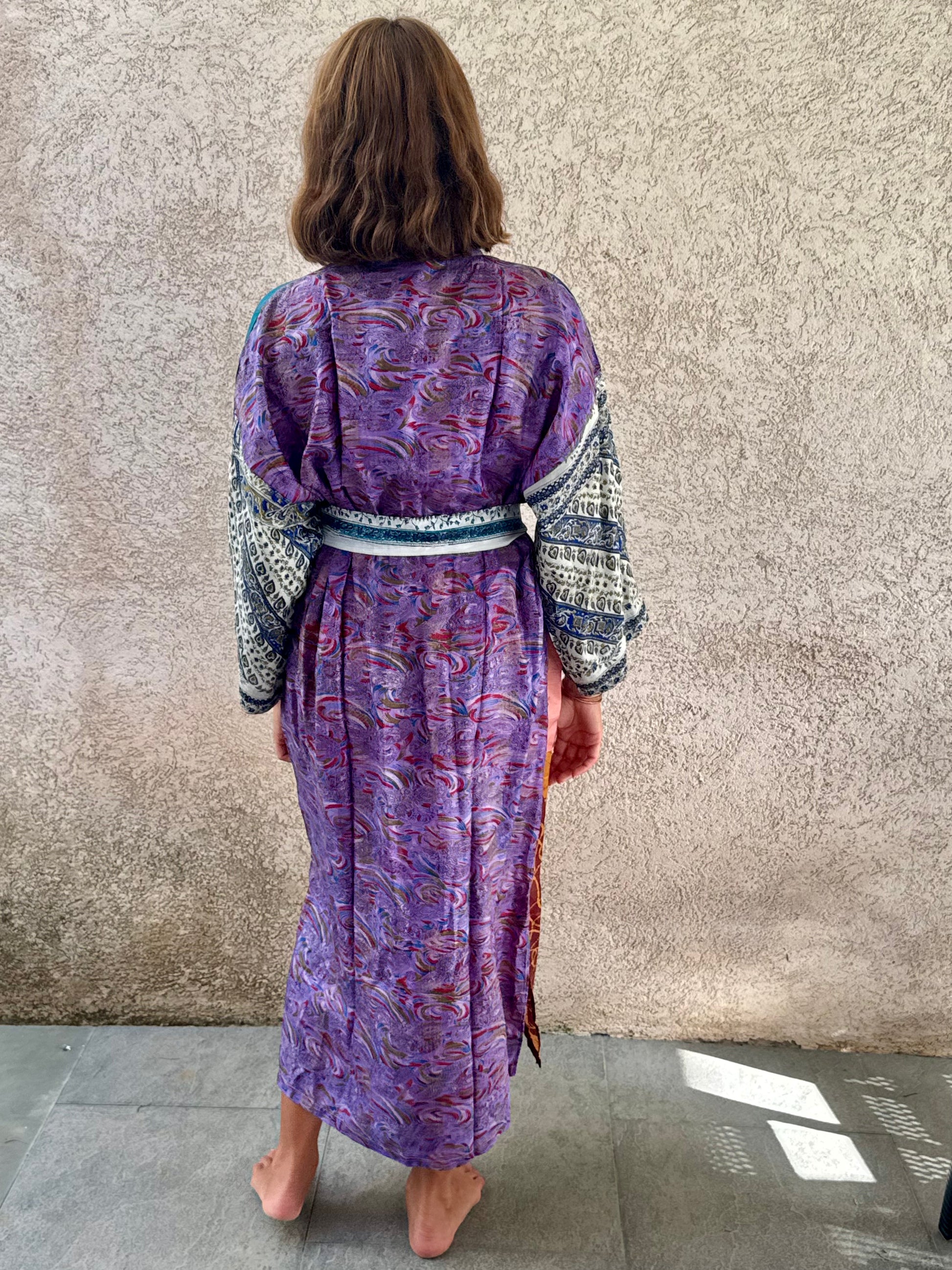 A woman viewed from behind wearing a purple patterned kaftan robe with patterned sleeves and a matching belt, standing barefoot against a textured concrete wall.